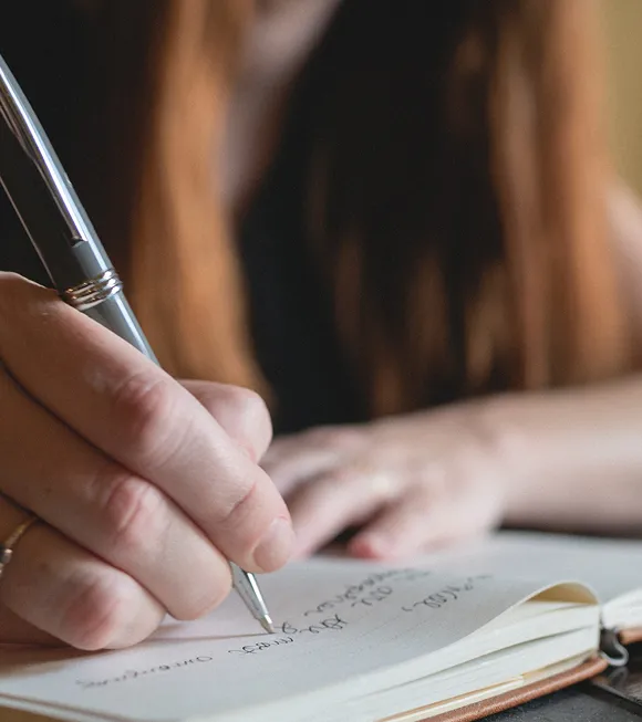 A woman focused on writing in a notebook, surrounded by a cozy, well-lit workspace.