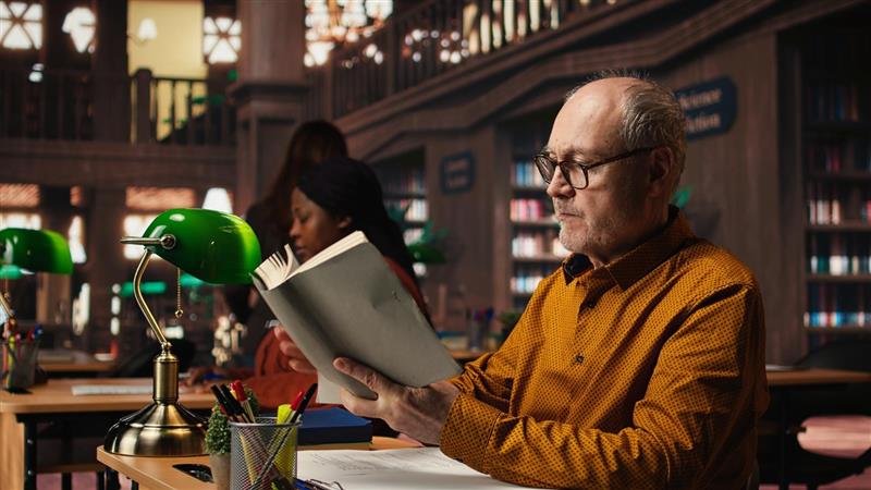 An older man reading a book in a quiet library, surrounded by shelves filled with books.